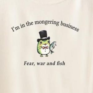 May include: A cream-coloured t-shirt featuring the phrase "I'm in the mongering business" and a cartoon frog in a top hat and bow tie, holding a fish. The text "Fear, war and fish" is printed below the frog.