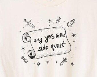 May include: Cream-colored t-shirt with rolled sleeves and a knotted hem. The shirt features a scroll design with the text "say yes to the side quest" surrounded by small illustrations of swords, potions, and stars.
