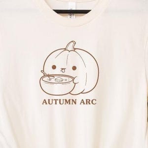 May include: A cream-coloured t-shirt with a cartoon pumpkin holding a bowl of soup. The pumpkin is outlined in brown and has a friendly face. The text "AUTUMN ARC" is printed below the pumpkin in brown.