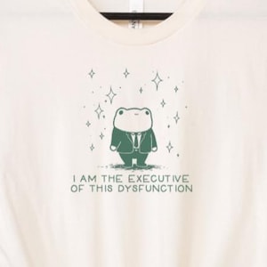 May include: Off-white t-shirt with a cartoon frog wearing a suit and tie. The frog is surrounded by stars, and the text "I AM THE EXECUTIVE OF THIS DYSFUNCTION" is printed below. The shirt is displayed on a black hanger.