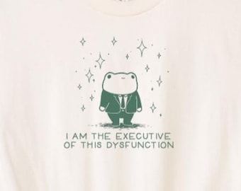 Può includere: T-shirt color crema con un ranocchio dei cartoni animati in giacca e cravatta. La rana è circondata da stelle e il testo "I AM THE EXECUTIVE OF THIS DYSFUNCTION" è stampato sotto. La maglietta è appesa a una gruccia nera.