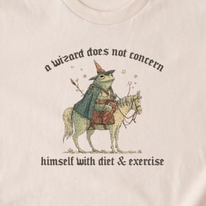 Puede incluir: Camiseta beige con una ilustración humorística de un mago rana a caballo. El texto dice "a wizard does not concern himself with diet & exercise". El diseño utiliza tonos verdes, marrones y beige.