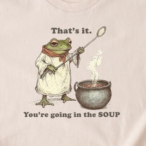 Puede incluir: Camiseta beige con un dibujo de una rana de dibujos animados vestida de chef, removiendo una olla de sopa humeante. El texto dice "That's it." y debajo, "You're going in the SOUP."
