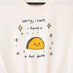 May include: Cream-colored t-shirt with a humorous graphic. The design features a smiling taco with the text "sorry, i can't, i have a hot date" in black. The design is surrounded by small diamond shapes.