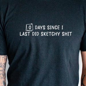May include: Black t-shirt with white text that reads "0 DAYS SINCE I LAST DID SKETCHY SHIT". The shirt is short-sleeved and has a crew neck. The person wearing the shirt has tattoos on their left arm.