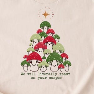 May include: Beige t-shirt featuring a whimsical design of a mushroom tree with red and green caps, and the text "We will literally feast on your corpse." A star is above the tree. The brand name "BOOOTEES" is at the bottom right.