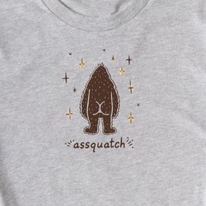 May include: A light gray t-shirt featuring a brown cartoon illustration of a Bigfoot from the rear, with the text "assquatch" below. The design includes star-like accents. The shirt has short sleeves and a crew neck.