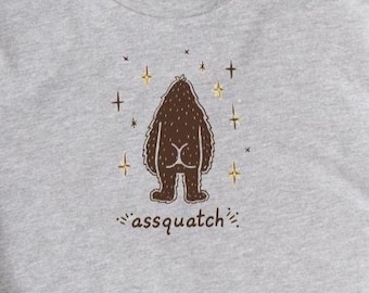Może przedstawiać: Jasnoszara koszulka z brązową, kreskówkową ilustracją Bigfoota od tyłu, z napisem "assquatch" poniżej. Projekt zawiera akcenty w kształcie gwiazd. Koszulka ma krótkie rękawy i okrągły dekolt.