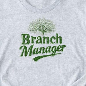 Puede incluir: Camiseta gris claro con la frase "Branch Manager" en verde y un diseño de árbol. La camiseta está anudada en la parte inferior y se encuentra sobre una alfombra redonda tejida.