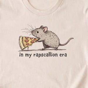 Op de afbeelding: Een beige t-shirt met een cartoonmuis die een pizza-punt vasthoudt. De tekst "in my rapscallion era" is onder de afbeelding gedrukt.