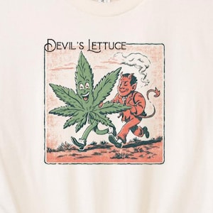May include: Cream-colored t-shirt featuring a cartoon graphic of a smiling cannabis leaf and a devil character running together. The text "Devil's Lettuce" is above the graphic. The shirt has short sleeves and a crew neck.