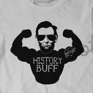 May include: A gray t-shirt with a black silhouette of Abraham Lincoln flexing his muscles. The text "HISTORY BUFF" is printed below the image. Lincoln has a tattoo on his arm that says "HISTORY BUFF".