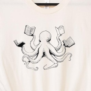 May include: Cream-colored t-shirt featuring a black and white illustration of an octopus holding books in its tentacles. The octopus is centered on the shirt, with the books open and visible. The shirt has short sleeves and a crew neck.