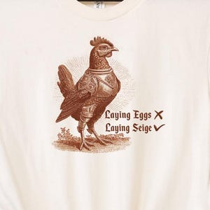 May include: Cream-colored t-shirt featuring a sepia-toned illustration of a chicken in armor. The text reads "Laying Eggs X, Laying Seige ✓". The shirt is hanging on a black hanger against a wooden background.