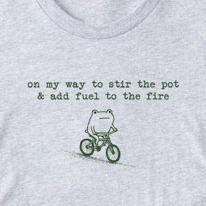 May include: A light gray t-shirt with the text "on my way to stir the pot & add fuel to the fire." A green frog rides a bicycle below the text. The shirt is made of a soft material.