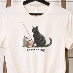 May include: A cream-coloured t-shirt with a black cat illustration. The cat is fishing with a rod and reel, with the word "catfishing" below. The shirt is hanging on a black hanger against a white background.