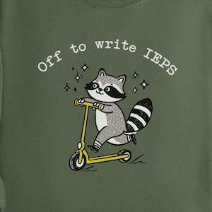 以下が含まれることがあります： 「Off to write IEPs」の文字と、黄色のスクーターに乗った漫画のアライグマが描かれたオリーブグリーンのスウェットシャツ。アライグマはグレーと白で、黒いマスクと縞模様の尻尾が付いています。スウェットシャツはクルーネックと長袖です。