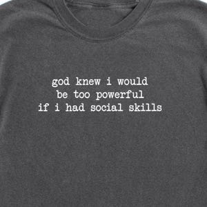 Pode incluir: T-shirt cinza escuro com o texto branco "god knew i would be too powerful if i had social skills". A camisa tem mangas enroladas e gola redonda. O nome da marca "booootees" é impresso na parte inferior.