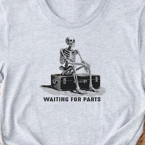 May include: A light gray t-shirt featuring a black and white illustration of a skeleton sitting on a trunk. The text "WAITING FOR PARTS" is printed below the image. The shirt has short sleeves and a crew neck.