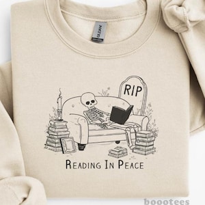 Może przedstawiać: Beżowa bluza z czarno-białą grafiką przedstawiającą szkielet czytający na kanapie, z nagrobkiem z napisem "RIP". Pod grafiką znajduje się napis "Reading in Peace". Bluza ma długie rękawy i okrągły dekolt.