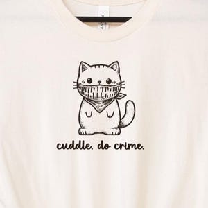 May include: Cream-colored t-shirt featuring a black and white cartoon cat wearing a bandana. The text "cuddle. do crime." is printed below the cat illustration. The shirt has short sleeves and a crew neck.