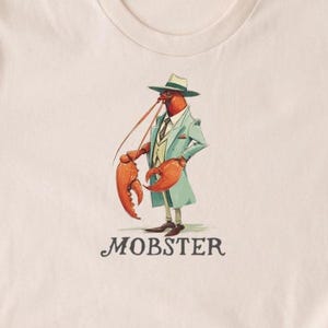 Puede incluir: Camiseta beige con un dibujo de una langosta de dibujos animados vestida con traje y sombrero, con la palabra "MOBSTER" debajo. La langosta lleva un traje verde claro y sostiene su pinza. La camiseta es de material suave.