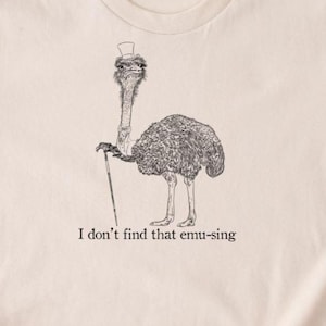 May include: Beige t-shirt with a black and white illustration of an ostrich wearing a top hat and holding a cane. The text "I don't find that emu-sing" is below the image. The shirt has short sleeves and a crew neck.