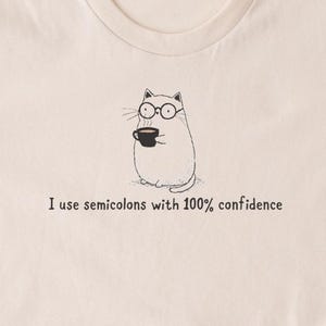 May include: A cream-coloured t-shirt with a cartoon cat wearing glasses and holding a coffee cup. The text "I use semicolons with 100% confidence" is printed below the cat illustration. The shirt has short sleeves and a crew neck.