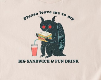 Może przedstawiać: Beżowa koszulka z kreskówkowym potworem jedzącym kanapkę i pijącym napój. Tekst brzmi: "Please leave me to my BIG SANDWICH & FUN DRINK." Potwór jest czarny z czerwonymi oczami i skrzydłami. Koszulka ma okrągły dekolt.