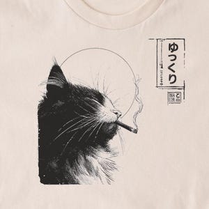 Può includere: T-shirt beige con un'illustrazione in bianco e nero di un gatto che fuma una sigaretta. La testa del gatto è di profilo, con fumo che sale. Testo giapponese stampato sul lato destro dell'immagine.