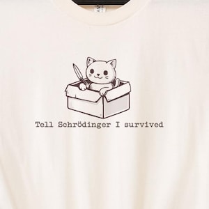 May include: Cream-colored t-shirt featuring a cartoon cat holding a knife while sitting in a box. The text "Tell Schrödinger I survived" is printed below the graphic. The shirt is hanging on a wooden ladder.