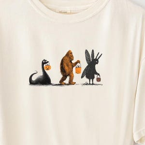 May include: Cream-colored t-shirt featuring a Halloween-themed graphic. The design depicts the Loch Ness Monster, Bigfoot, and a creature with rabbit ears, each holding a trick-or-treat bucket. The shirt has a relaxed fit and a crew neck.