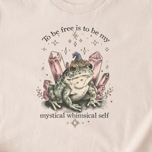 May include: A light beige t-shirt featuring a whimsical illustration of a frog wearing a hat, surrounded by crystals. The text "To be free is to be my mystical whimsical self" is printed above and below the frog. The shirt has short sleeves.