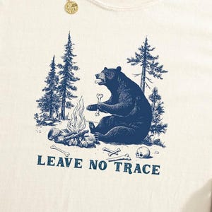 May include: Cream-coloured t-shirt featuring a blue graphic of a bear by a campfire, with trees. The text "LEAVE NO TRACE" is printed below. The shirt is made of a soft material.