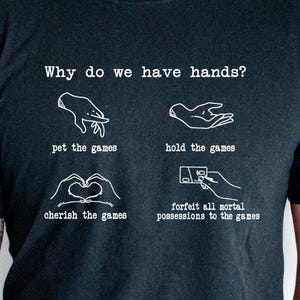 May include: Black t-shirt with white text and illustrations. The text reads "Why do we have hands?" followed by four phrases: "pet the games," "hold the games," "cherish the games," and "forfeit all mortal possessions to the games."
