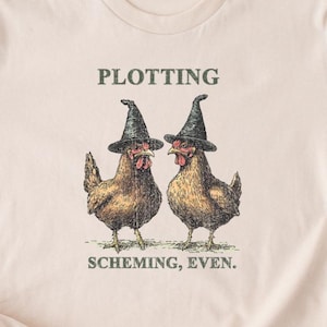 May include: A beige t-shirt with a graphic of two chickens wearing witch hats. The text "Plotting Scheming, Even." is printed below the chickens.
