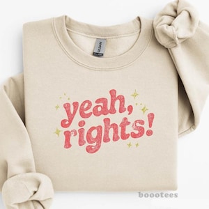 May include: A beige sweatshirt with the phrase "yeah, rights!" in red, retro-style lettering. The design includes small gold stars. The sweatshirt is folded neatly on a white surface.