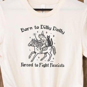May include: Cream-colored t-shirt featuring a black graphic of a frog riding a unicorn, accompanied by a black cat. The text reads "Born to Dilly Dally" above and "Forced to Fight Fascists" below the illustration. The shirt is knotted at the bottom.