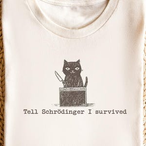 Puede incluir: Camiseta color crema con una ilustración de un gato negro sosteniendo un cuchillo, sentado en una caja. El texto "Tell Schrödinger I survived" está impreso debajo. La camiseta se muestra sobre una superficie beige tejida.