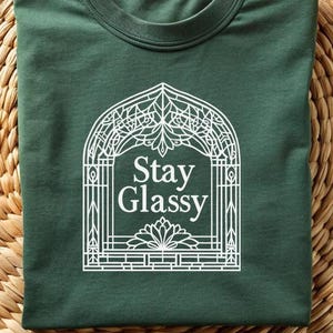 May include: Dark green t-shirt with the text "Stay Glassy" in a white decorative frame. The shirt is folded on a woven, circular surface. The design features floral and geometric elements.