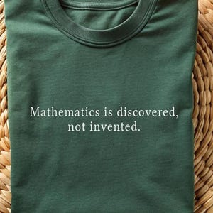 Pode incluir: T-shirt verde escuro com o texto "A matemática é descoberta, não inventada". A camisa está dobrada cuidadosamente sobre uma superfície tecida marrom claro. A camisa tem gola redonda e mangas curtas.