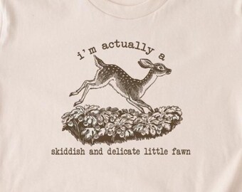 Op de afbeelding: Beige t-shirt met een afbeelding van een rennend hert boven een bloembed. De tekst luidt: "I'm actually a skiddish and delicate little fawn". De merknaam "BOOOTEES" staat onderaan.