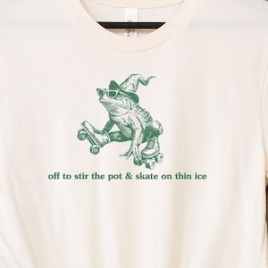 May include: Cream-colored t-shirt featuring a green graphic of a frog wearing sunglasses and a witch hat, riding roller skates. The text "off to stir the pot & skate on thin ice" is printed below the graphic. The shirt has rolled-up sleeves.