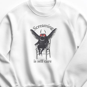 May include: White sweatshirt with a black and white illustration of a creature with wings and red eyes sitting on a chair. The text "Screaming is self care" is printed below the illustration.