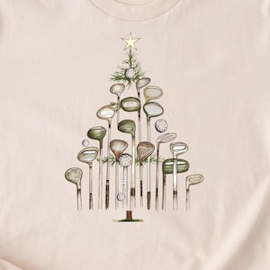 Puede incluir: Camiseta beige claro con un diseño de árbol de Navidad hecho con palos de golf. El árbol está adornado con una estrella, pelotas de golf y agujas de pino. El diseño utiliza tonos verdes, marrones y blancos.