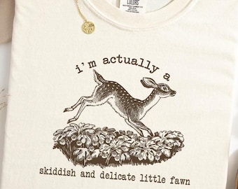 Op de afbeelding: Crèmekleurig T-shirt met een bruine afbeelding van een springend hert en de tekst "i'm actually a skiddish and delicate little fawn". Het shirt maakt deel uit van de Comfort Colors collectie.