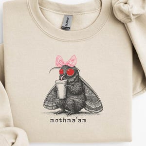 Puede incluir: Sudadera beige con una ilustración en blanco y negro de una polilla con un lazo rosa y gafas de sol rojas, sosteniendo una bebida. El texto "mothma'am" está impreso debajo. La sudadera tiene cuello redondo y mangas largas.