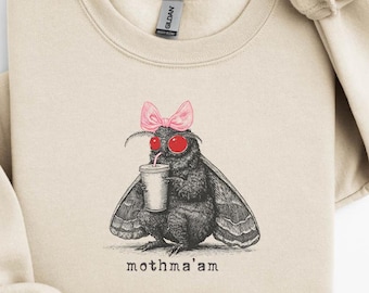 May include: Beige sweatshirt featuring a black and white illustration of a moth wearing a pink bow and red sunglasses, holding a drink. The text "mothma'am" is printed below the moth. The sweatshirt has a crew neck and long sleeves.
