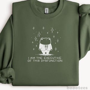 Può includere: Felpa verde oliva con grafica bianca di una rana in giacca e cravatta, circondata da stelle. Il testo "I AM THE EXECUTIVE OF THIS DYSFUNCTION" è stampato sotto. Il marchio "boootees" è stampato in basso a destra.
