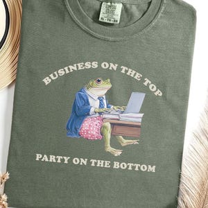 Könnte beinhalten: Olivgrünes T-Shirt mit dem Text "BUSINESS ON THE TOP PARTY ON THE BOTTOM". Die Grafik zeigt einen Frosch im Anzug, der an einem Laptop arbeitet und rosa gepunktete Shorts trägt. Ein Strohhut und eine Feder sind im Hintergrund.